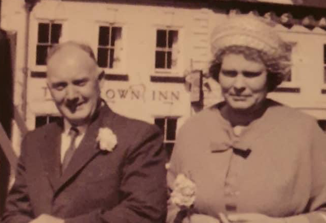 A sepia-toned photograph of two people, a man and a woman, standing in front of a British public house called The Crown Inn.