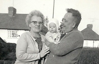 A black-and-white photograph of a woman and a man standing in their front garden, the man is holding a small child.